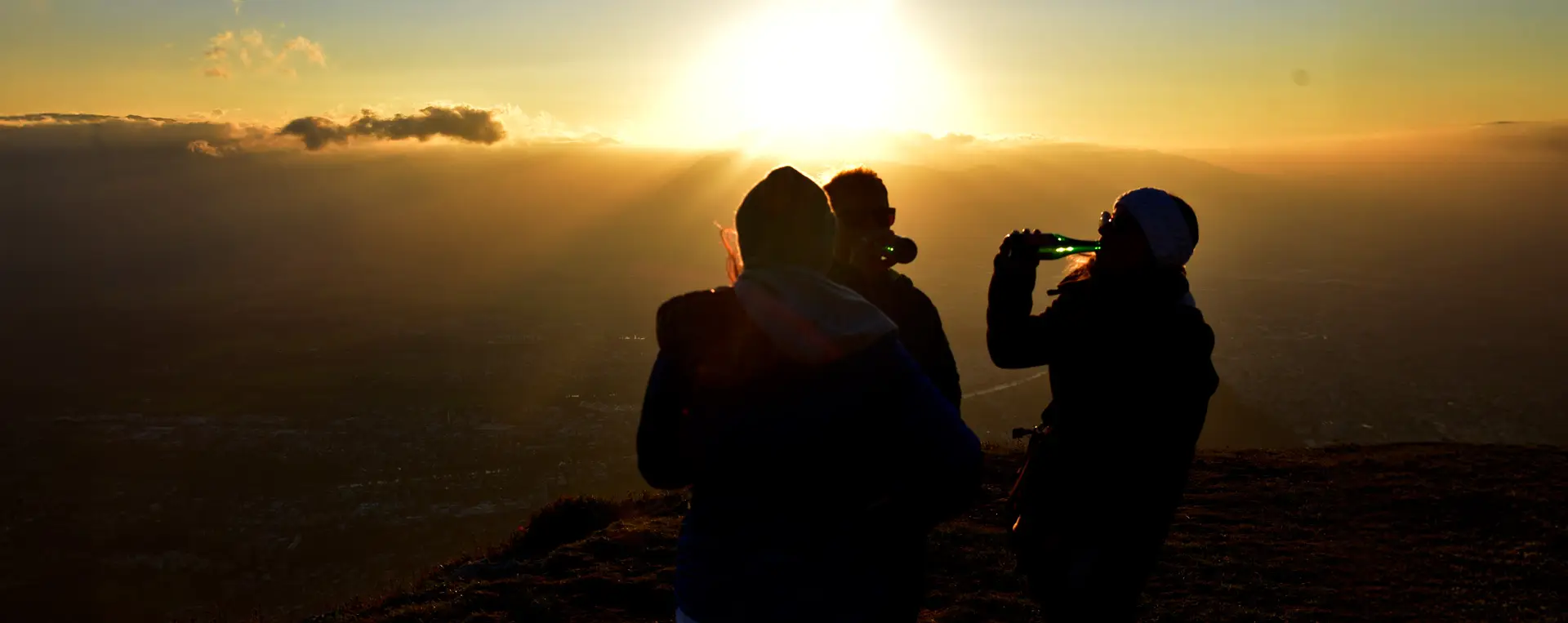 Three people enjoying local beers on top of Gaisberg during the Salzburg Night Tour, with stunning views and a bright sun setting the atmosphere.