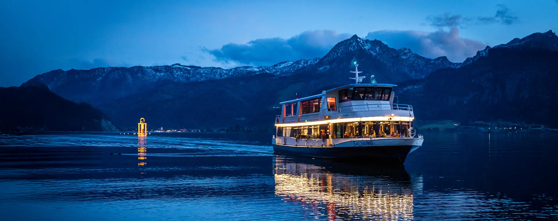 A ship on Lake Wolfgang at night, surrounded by towering mountains with a distant Christmas market glowing in the background.