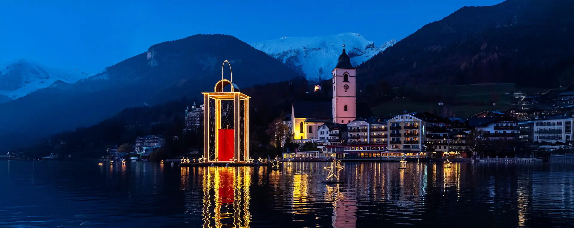 Illuminated floating lantern near St. Wolfgang, with the Weißes Rössl and snow-covered Schafberg in the backdrop.